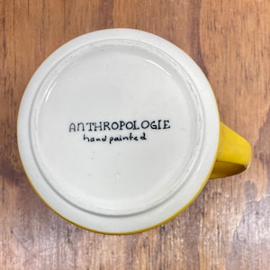 Peut inclure: Une tasse en c&eacute;ramique jaune et blanche avec une anse. Le fond de la tasse porte l'inscription "ANTHROPOLOGIE hand painted" en noir. La tasse est pos&eacute;e sur une surface en bois.