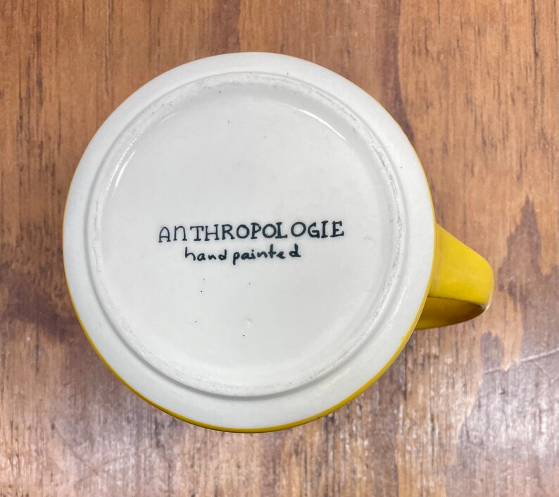 Peut inclure: Une tasse en c&eacute;ramique jaune et blanche avec une anse. Le fond de la tasse porte l'inscription "ANTHROPOLOGIE hand painted" en noir. La tasse est pos&eacute;e sur une surface en bois.