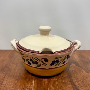 May include: Cream-colored ceramic soup bowl with a matching lid. The bowl features a yellow base, a decorative band with olive branch designs, and a dark red rim. It has two handles and a small opening in the lid for a spoon.