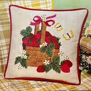 May include: A square throw pillow cover featuring an embroidered basket filled with red strawberries, green leaves, and white flowers. A pink ribbon is tied to the basket handle, and two yellow butterflies are embroidered nearby. The pillow has red piping.