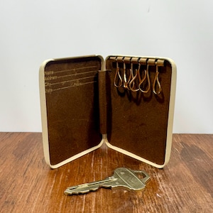 May include: Open, cream key case with a brown interior and gold key hooks. A gold key rests on the wooden surface in front of the open case. The interior has space for name, address, city, and state.
