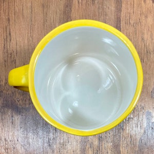 Peut inclure: Une tasse &agrave; caf&eacute; jaune avec un int&eacute;rieur blanc. La tasse a une anse arrondie et un bord jaune vif. La tasse est vide et repose sur une surface en bois.
