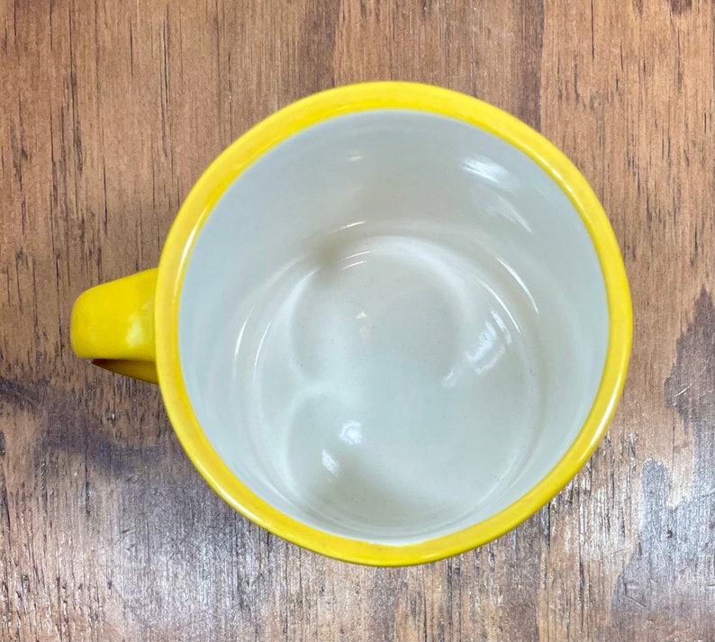 Peut inclure: Une tasse &agrave; caf&eacute; jaune avec un int&eacute;rieur blanc. La tasse a une anse arrondie et un bord jaune vif. La tasse est vide et repose sur une surface en bois.