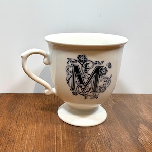 Puede incluir: Taza de cer&aacute;mica color crema con base de pedestal y asa curva. La taza presenta una letra "M" negra adornada con detalles florales. La taza est&aacute; sobre una superficie de madera.