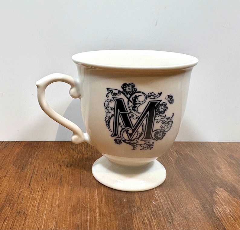 Puede incluir: Taza de cer&aacute;mica color crema con base de pedestal y asa curva. La taza presenta una letra "M" negra adornada con detalles florales. La taza est&aacute; sobre una superficie de madera.