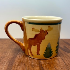 May include: A ceramic coffee mug with a tan background and a brown moose illustration. The mug has a brown handle and a green stripe near the rim. Two green trees are also painted on the mug.