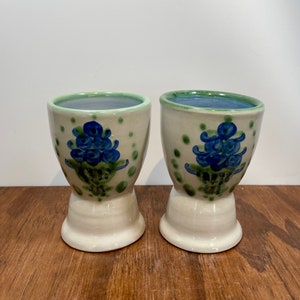 May include: Two ceramic egg cups. The cups are off-white with a blue floral design and green polka dots. The interior is blue, and the rim is green. The cups sit on a white pedestal base.