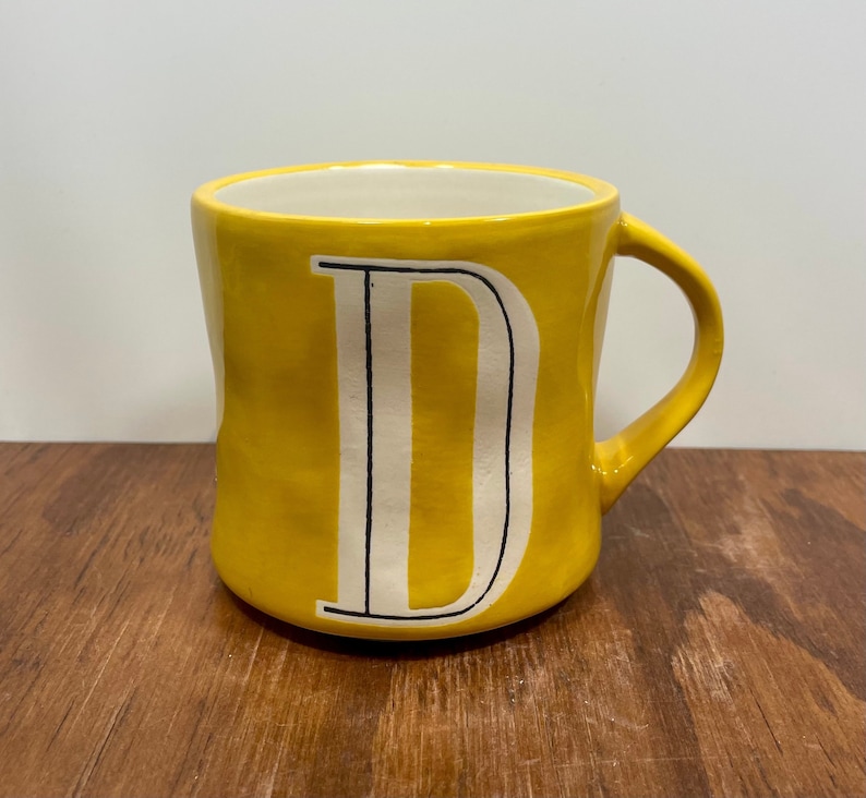 Peut inclure: Une tasse en c&eacute;ramique jaune avec un int&eacute;rieur blanc et une grande lettre "D" blanche avec des contours noirs. La tasse a une forme arrondie et une anse incurv&eacute;e. La tasse est pos&eacute;e sur une surface en bois.
