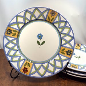 May include: A set of white ceramic plates with a floral design. Each plate features a blue flower in the centre and a border with blue, green, and yellow geometric patterns. Additional plates are stacked in the background.