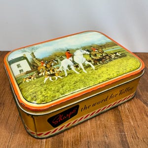 May include: A rectangular metal tin with rounded corners featuring a colorful illustration of a fox hunt on the lid. The side of the gold-colored container displays the text Sharps the word for Toffee in black and red script with diagonal stripes.