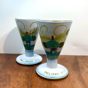 May include: Two white ceramic goblets with a flared top and a pedestal base. Each goblet is decorated with green and blue floral designs and the words "PELICAN HILL" in green lettering on the base. The goblets are on a wooden surface.
