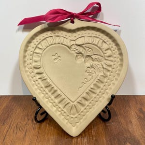 May include: A heart-shaped beige ceramic mold featuring a raised embossed design of a small bee, a flower, and leaves. The border has a ruffled texture, and a bright red satin ribbon is tied through a hole at the top for hanging purposes.