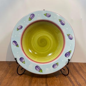 Könnte beinhalten: Dekorative Keramik-Servierschüssel mit hellblauem Rand, verziert mit lila Auberginen-Illustrationen und weißen Punkten. Die Schüssel hat ein grün-gelbes Spiralmuster in der Mitte und einen rosa Innenrand. Die Schüssel wird auf einem schwarzen Metallständer präsentiert.