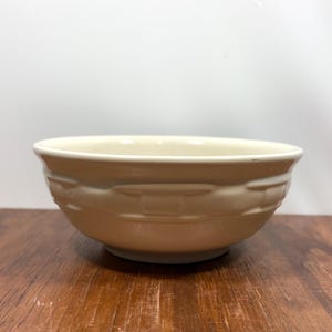 Puede incluir: Un cuenco de cerámica beige con un diseño de tejido de cesta en el exterior. El cuenco tiene forma redondeada y está sobre una superficie de madera. El interior del cuenco es de un tono beige más claro. Diámetro: unos 20 cm.
