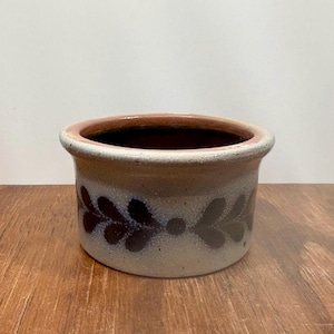 Puede incluir: Un pequeño cuenco redondo de gres con un cuerpo beige claro y un interior marrón oscuro. El borde es de un marrón más oscuro. El exterior presenta un patrón repetido de hojas marrones oscuras. El cuenco está sobre una superficie de madera.