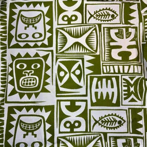 May include: A fabric textile featuring a repeating block print pattern in olive green and white. The design consists of various rectangular panels containing stylised graphic motifs, including faces, fish skeletons, and abstract geometric shapes.