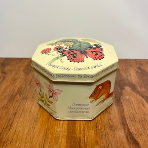 May include: A cream-colored, octagonal tin with illustrations of a butterfly and a dormouse. The lid features a Painted Lady butterfly and text that reads "The Countryside Collection by Bristows - Rural Life". The side of the tin has a dormouse illustration and the scientific name "Muscardinus avellanarius".