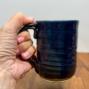 May include: A handmade ceramic mug with a dark blue glaze and a textured, ridged design. The mug has a dark brown handle and base, and a light-colored bottom. The mug is held in a hand.