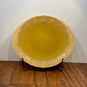 Puede incluir: Una fuente ovalada de color mostaza con un borde decorativo. El borde presenta un patrón repetitivo de motivos florales y de urnas, con una textura similar a una cuerda. La fuente se exhibe sobre un soporte de metal negro.