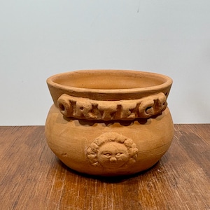 以下が含まれることがあります： テラコッタ色の植木鉢。広い縁と丸みを帯びたボディが特徴です。彫刻された文字と太陽のような顔が施された装飾的なバンドがあります。屋内または屋外での使用に適しています。
