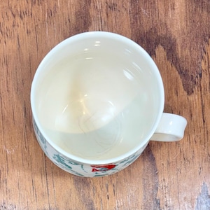 K&ouml;nnte beinhalten: Wei&szlig;er Keramikbecher mit Henkel, mit einem dekorativen Muster aus gr&uuml;nen und roten Akzenten. Der Becher ist leer und steht auf einer Holzoberfl&auml;che. Das Innere des Bechers ist hellcremefarben.