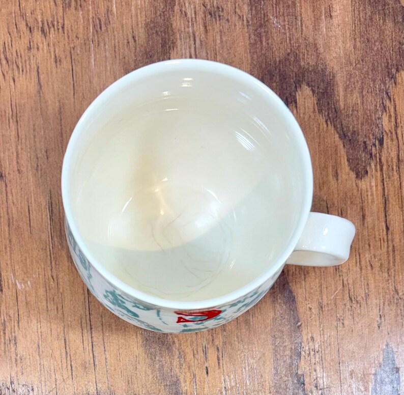 K&ouml;nnte beinhalten: Wei&szlig;er Keramikbecher mit Henkel, mit einem dekorativen Muster aus gr&uuml;nen und roten Akzenten. Der Becher ist leer und steht auf einer Holzoberfl&auml;che. Das Innere des Bechers ist hellcremefarben.