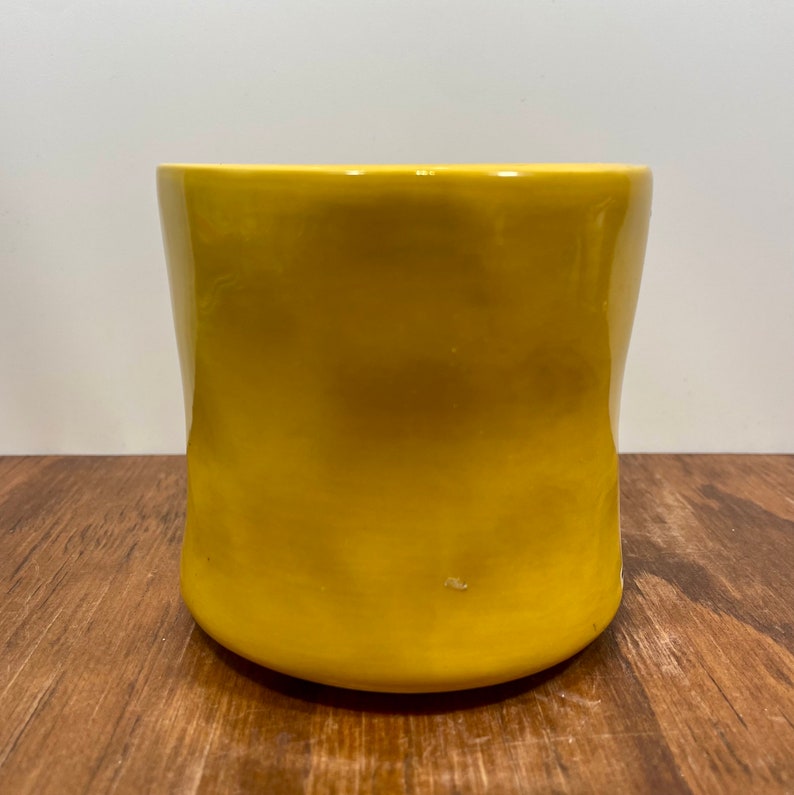 Peut inclure: Un pot de fleur en c&eacute;ramique jaune avec une finition brillante. Le pot cylindrique a une forme l&eacute;g&egrave;rement incurv&eacute;e et repose sur une surface en bois. Le pot est vide et pr&ecirc;t pour une plante.