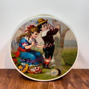 May include: A round metal tin featuring a vintage illustration of a person in a blue dress sewing a button onto a brown jacket worn by another person. The scene includes a basket of flowers, a straw hat, and a spool of Clark's O.N.T. thread.