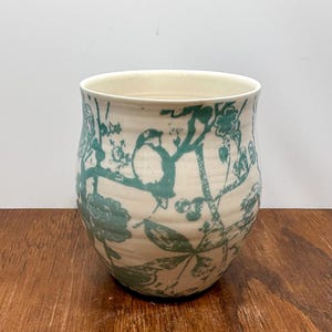 K&ouml;nnte beinhalten: Eine cremefarbene Keramikvase mit einem tealfarbenen Vogel- und Blumendesign. Die Vase hat eine weite &Ouml;ffnung und eine leicht geschwungene Form. Das Design umfasst V&ouml;gel, Bl&auml;tter und Blumen. Die Vase steht auf einer Holzoberfl&auml;che.