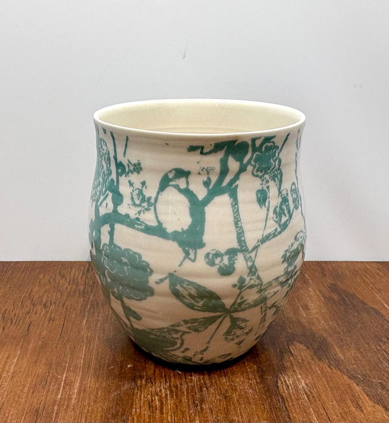K&ouml;nnte beinhalten: Eine cremefarbene Keramikvase mit einem tealfarbenen Vogel- und Blumendesign. Die Vase hat eine weite &Ouml;ffnung und eine leicht geschwungene Form. Das Design umfasst V&ouml;gel, Bl&auml;tter und Blumen. Die Vase steht auf einer Holzoberfl&auml;che.