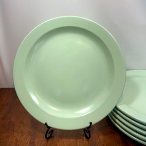 Puede incluir: Un conjunto de platos verde claro. Los platos son redondos con un borde ligeramente elevado. Los platos se exhiben en un soporte de metal negro. Los platos tienen un acabado liso y brillante. Los platos son de color verde pastel.