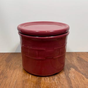 May include: A burgundy ceramic jar with a matching lid. The jar has a cylindrical shape with a textured, basket-weave design around the body. The lid sits flush on top. The jar is on a wooden surface.