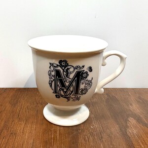 Puede incluir: Taza de cer&aacute;mica color crema con la letra "M" negra y dise&ntilde;o floral. La taza tiene una base de pedestal y un asa curva. El dise&ntilde;o est&aacute; centrado en el cuerpo de la taza. La taza est&aacute; sobre una superficie de madera.