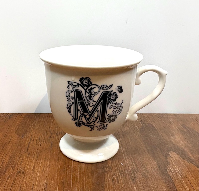 Puede incluir: Taza de cer&aacute;mica color crema con la letra "M" negra y dise&ntilde;o floral. La taza tiene una base de pedestal y un asa curva. El dise&ntilde;o est&aacute; centrado en el cuerpo de la taza. La taza est&aacute; sobre una superficie de madera.