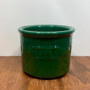 May include: A glossy, emerald green ceramic pot with a rounded rim and horizontal embossed bands. The pot sits on a wooden surface, offering a rustic contrast to the vibrant green. This decorative pot is suitable for plants or storage.