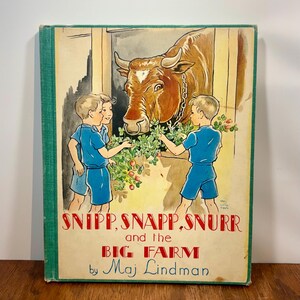 Puede incluir: Un libro de tapa dura vintage titulado Snipp, Snapp, Snurr and the Big Farm de Maj Lindman. La portada muestra un toro marrón en un granero con tres niños vestidos de azul alimentándolo con follaje verde y bayas rojas. Lomo verde.