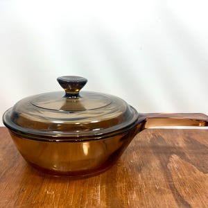 Puede incluir: Una cacerola de vidrio ámbar vintage con tapa a juego. La cacerola tiene un mango largo y una forma redondeada. La tapa tiene un pequeño pomo oscuro. La batería de cocina está hecha de vidrio transparente.