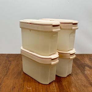 May include: Four beige plastic food storage containers with light tan lids are stacked on a wooden surface. The containers are rectangular with rounded corners. The lids have a curved design and a small handle on each side.
