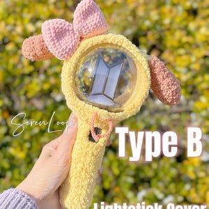 May include: A yellow lightstick cover with a pink bow and ear-like accents. The cover has the text "Type B Lightstick Cover" in white. The lightstick is held in a hand.