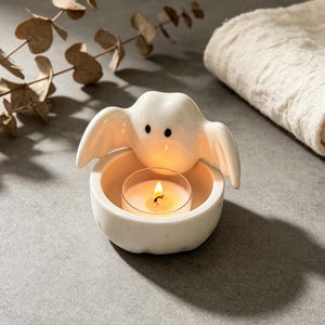 May include: A white ceramic ghost-shaped tea light candle holder with two small black eyes. The ghost figure is positioned behind a circular base that holds a single lit tea light candle with a visible yellow flame and a clear plastic cup.