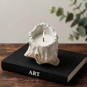 May include: A white, textured candle holder shaped like a tree stump with a lit white candle inside. The candle holder is resting on a black book with the word ART printed in white letters on the spine, set against a neutral background.