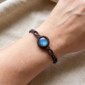 Peut inclure: Un bracelet en macramé marron orné d'une pierre précieuse bleue irisée et ronde. Le bracelet est tissé de manière complexe avec une série de nœuds et de petits accents métalliques. La pierre précieuse est le point central, affichant une gamme de teintes bleues.