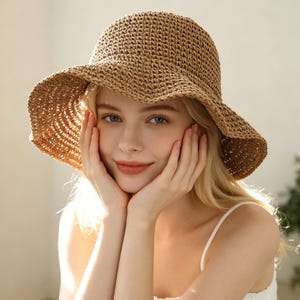 May include: A tan, woven straw bucket hat with a wide, flexible brim. The hat features a textured, crocheted pattern throughout the crown and brim, providing a structured yet pliable shape that frames the head with a natural, earthy tone.