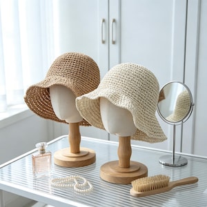 May include: Two crochet bucket hats, one in tan and one in cream, displayed on wooden mannequin heads. The hats feature a wide, wavy brim and an open-weave texture. A glass perfume bottle, a pearl necklace, and a wooden hairbrush are nearby.