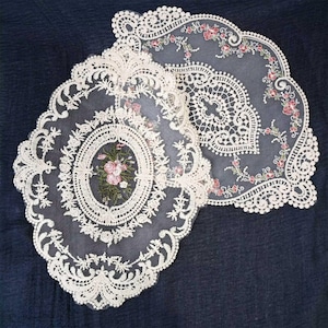 May include: Two off-white lace doilies with floral embroidery. One is oval-shaped with a central floral design, the other is a scalloped shape with floral accents. The doilies are set against a dark blue background.