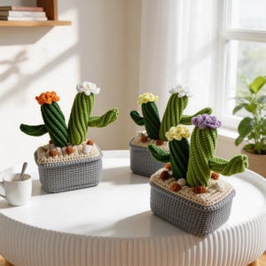May include: Four handmade crochet cactus plants in gray rectangular pots. The cacti are green with various colored flower accents, including orange, white, yellow, and purple. The pots have a light tan base with small brown decorative stones.
