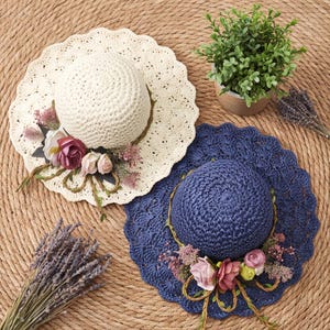 May include: Two crocheted sun hats, one cream and one navy blue, each featuring a scalloped brim and a decorative arrangement of artificial pink and white roses with green foliage and thin brown twine accents attached to the front.
