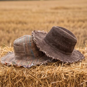 Può includere: Due cappelli in stile fedora con bordi sfrangiati. Uno è di colore marrone scuro, mentre l'altro presenta un motivo intrecciato multicolore con striature blu, arancioni e beige. Entrambi i cappelli hanno una corona strutturata e una tesa larga.