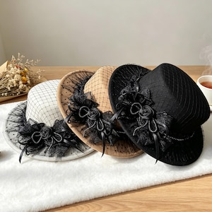 Puede incluir: Tres sombreros en blanco, beige y negro, cada uno con una copa estructurada y ala ancha. Los sombreros están adornados con un velo de red negra, un lazo de cinta negra decorativo y una rosa de tela negra en la parte frontal.