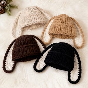 May include: Four chunky knit beanies with long, floppy rabbit ears. The hats come in cream, tan, dark brown, and black. Each beanie features a ribbed cuff and long, soft-textured ears with a contrasting white inner lining on the underside.